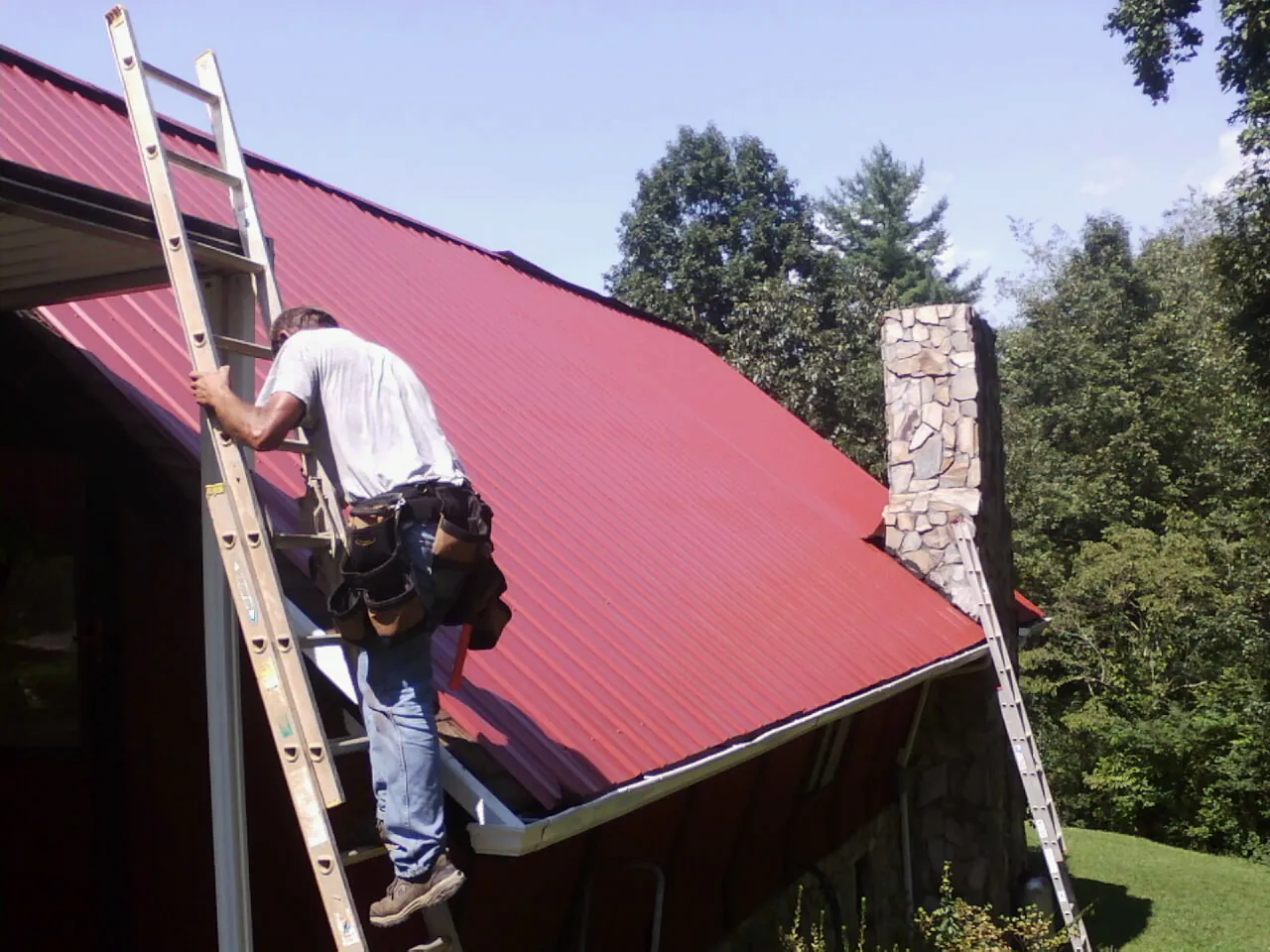 Roofer on a ladder beside a red metal roof and stone chimney — Soffit & Fascia Repair by Roofing Brothers in Lexington Park